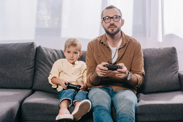 Padre y pequeño hijo sentado en el sofá y jugando con joysticks - foto de stock
