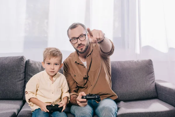 Padre e hijo pequeño sentados en el sofá y jugando con gamepads - foto de stock