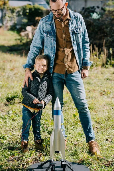 Счастливый отец с маленьким сыном, улыбающимся в камеру во время игры с моделью ракеты на открытом воздухе — стоковое фото