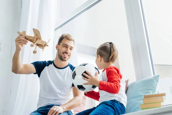 Junge mit Fußballball sieht lächelnden Vater mit Holzspielzeugflugzeug zu Hause an — Stockfoto