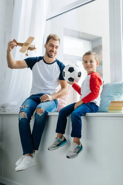 Niño sonriente con pelota de fútbol y padre con avión de juguete de madera en casa - foto de stock