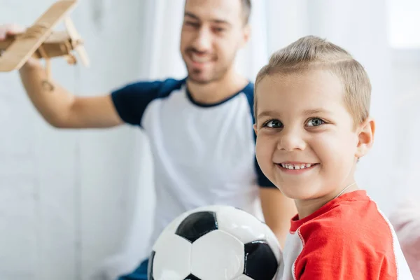 Foco seletivo de menino sorridente com bola de futebol e pai com avião de brinquedo de madeira em casa — Fotografia de Stock