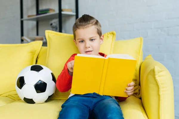 Портрет маленького мальчика, читающего книгу на жёлтом диване с футбольным мячом рядом дома — стоковое фото