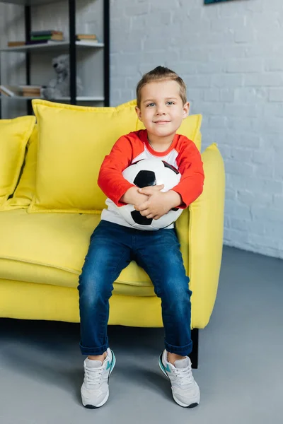 Lächelnder Junge mit Fußball in der Hand auf gelbem Sofa zu Hause — Stockfoto