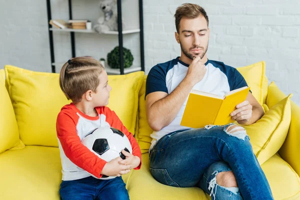 Bambino con pallone da calcio guardando il libro di lettura del padre sul divano a casa — Stock Photo
