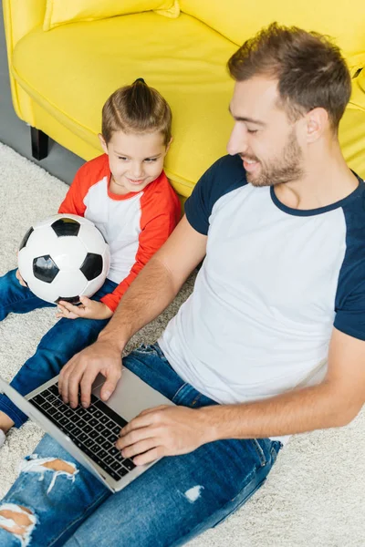 Hombre usando portátil mientras pequeño hijo con pelota de fútbol sentado cerca en el suelo en casa - foto de stock