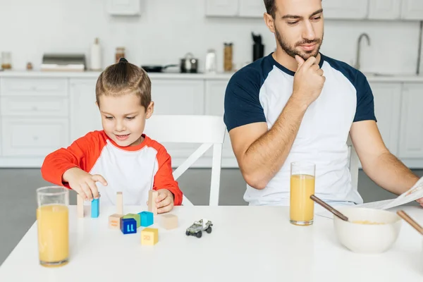 Homem lendo jornal enquanto filho brincando com cubos na mesa na cozinha — Fotografia de Stock