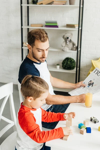 Man with newspaper looking at little son playing with wooden blocks at table in kitchen — Stock Photo