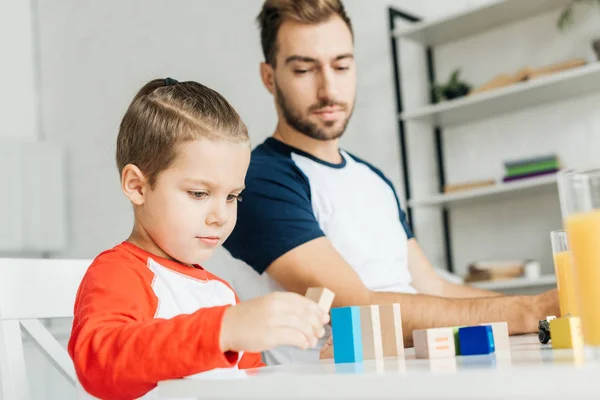 Man looking at little son playing with wooden blocks at table in kitchen — Stock Photo