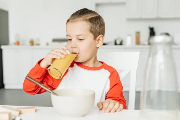 Menino bebendo suco de vidro enquanto toma café da manhã na cozinha em casa — Fotografia de Stock