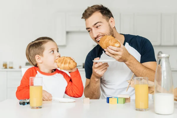 Joven padre e hijo desayunando con croissants y zumo de naranja juntos y mirándose en casa - foto de stock