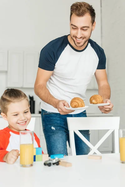 Красивий молодий батько, який подає круасани на сніданок з сином, граючи з іграшками — стокове фото