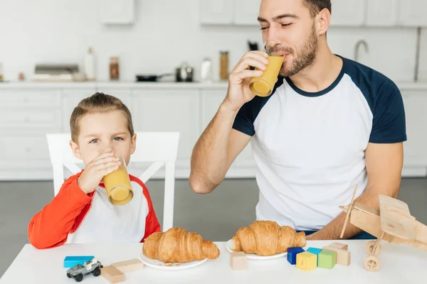 Jovem pai e filho tomando café da manhã com croissants e suco de laranja juntos em casa — Fotografia de Stock