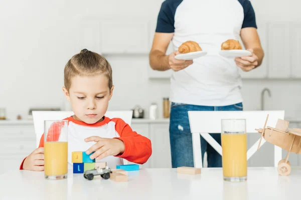 Cropped shot of father serving croissants for breakfast with son while he playing with toys — Stock Photo