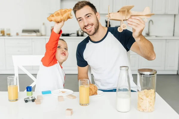 Hermoso joven padre e hijo jugando con croissant y juguete avión mientras desayunan juntos en casa - foto de stock