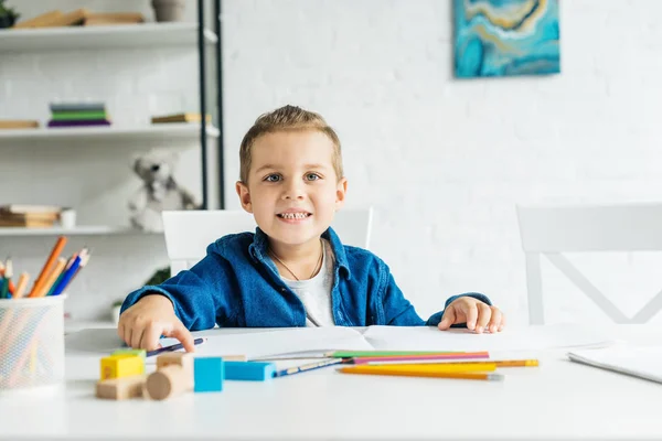 Petit enfant souriant dessinant avec des crayons de couleur à la maison et regardant la caméra — Photo de stock