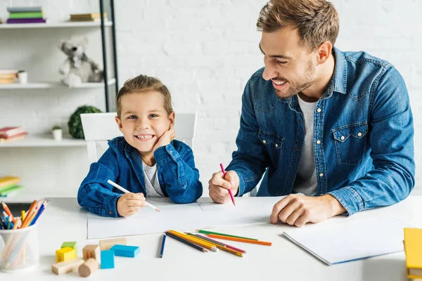 Красивый молодой отец рисует с улыбающимся маленьким сыном дома — стоковое фото