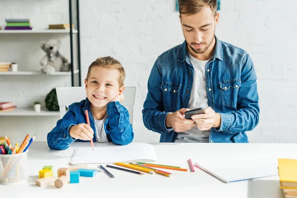 Beau jeune père en utilisant smartphone tandis que son fils dessin à la maison — Photo de stock