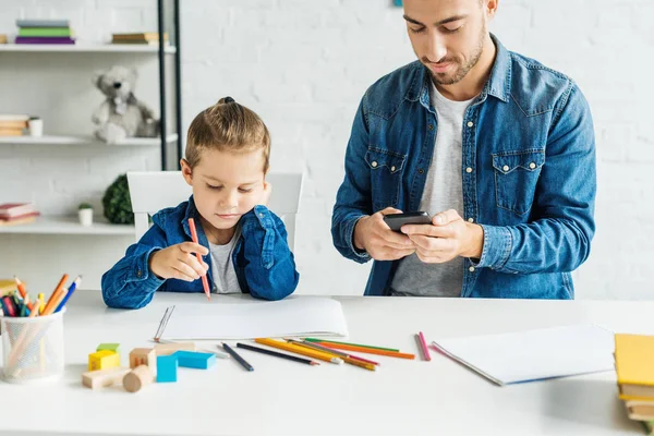 Giovane padre utilizzando lo smartphone mentre il figlio disegna a casa — Foto stock
