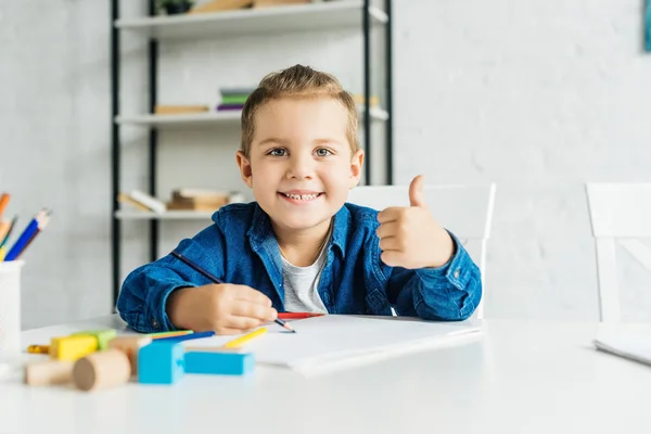 Счастливый малыш, показывающий большой палец, когда рисует дома — стоковое фото