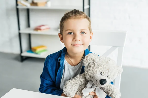 Entzückendes kleines Kind sitzt mit Teddybär am Tisch und blickt in die Kamera — Stockfoto