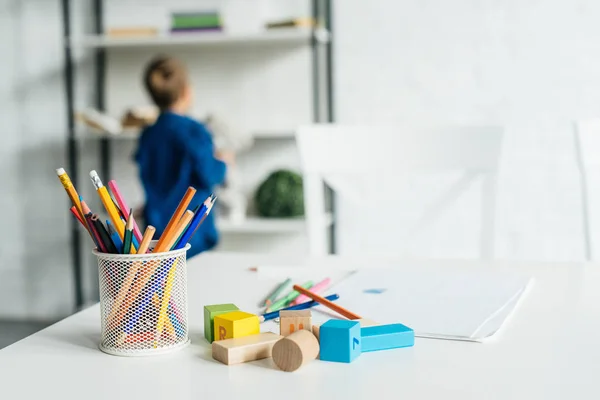 Lápices de color y bloques de madera en la mesa con cuaderno acostado en la mesa con un niño pequeño borroso en el fondo - foto de stock