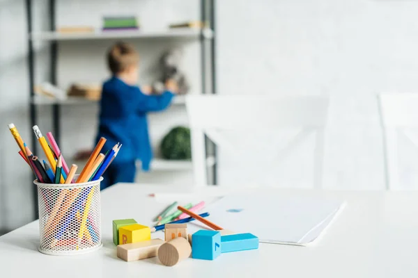 Farbstifte und Holzklötze auf dem Tisch mit Album auf dem Tisch liegend mit kleinem Kind auf dem Hintergrund verschwommen — Stockfoto