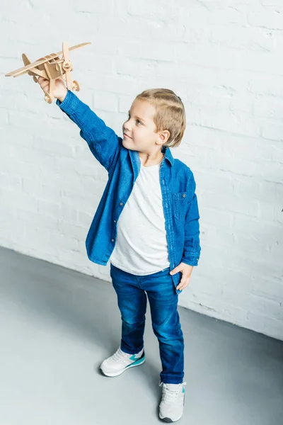 Adorable little kid playing with wooden toy airplane in front of white brick wall — Stock Photo