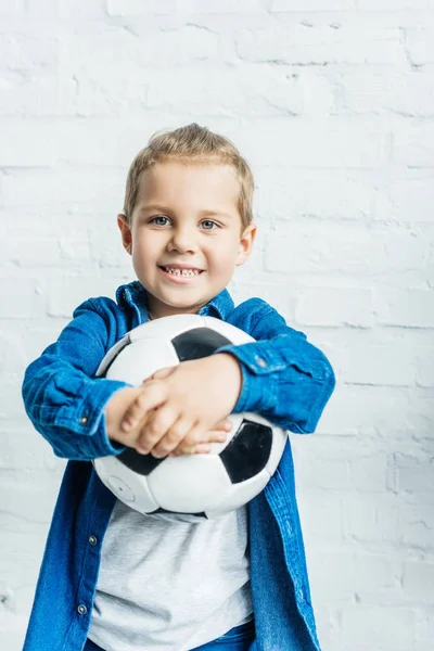 Niño feliz con pelota de fútbol mirando a la cámara delante de la pared de ladrillo blanco — Stock Photo