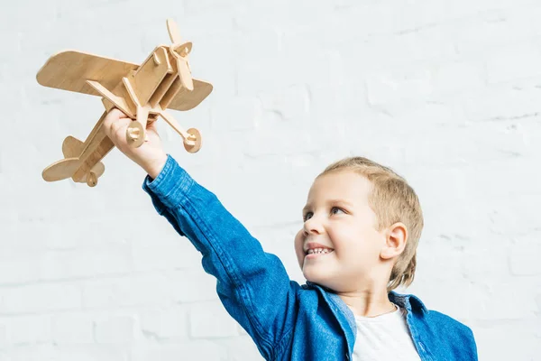 Criança sorrindo brincando com avião de brinquedo de madeira na frente da parede de tijolo branco — Fotografia de Stock