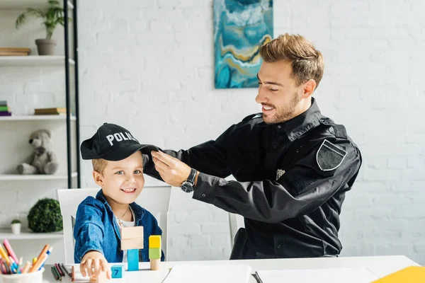 Beau jeune père en uniforme de police mettant casquette de police sur les fils tête à la maison — Photo de stock