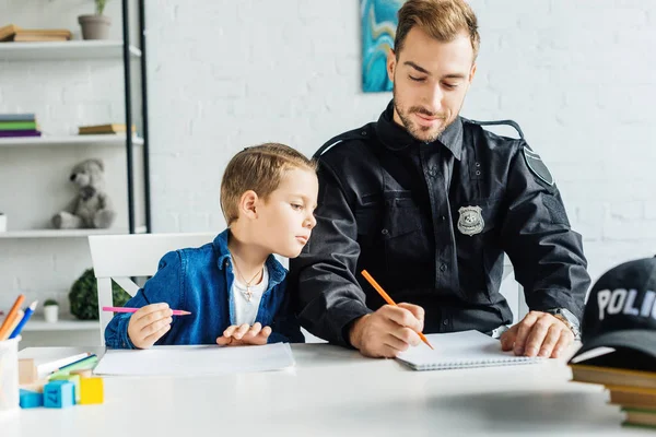 Красивый молодой отец в полицейской форме и сын рисуют вместе дома — стоковое фото