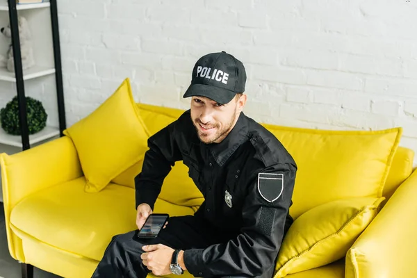 Glücklicher junger Polizist mit Smartphone sitzt auf gelbem Sofa und blickt in die Kamera — Stockfoto
