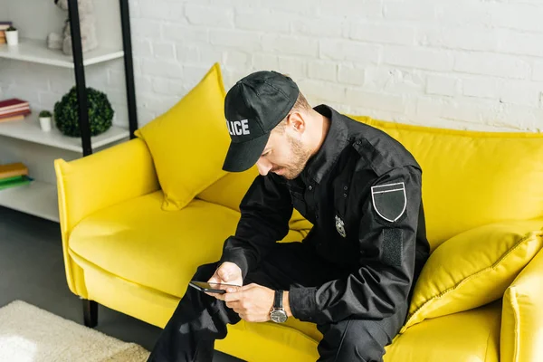 Bonito jovem policial com smartphone sentado no sofá amarelo — Fotografia de Stock