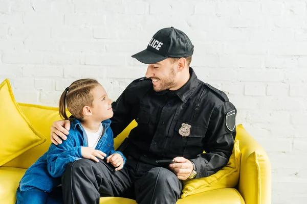 Guapo joven padre en uniforme de policía e hijo pasar tiempo juntos mientras están sentados en el sofá amarillo en casa - foto de stock