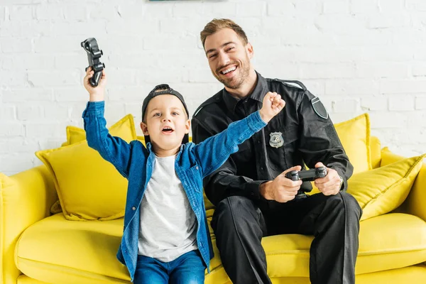 Bonito jovem pai no polícia uniforme e filho enquanto sentado no sofá amarelo em casa — Fotografia de Stock