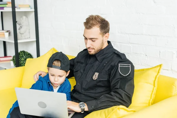 Beau jeune père en uniforme de police et fils utilisant ordinateur portable ensemble tout en étant assis sur le canapé jaune à la maison — Photo de stock