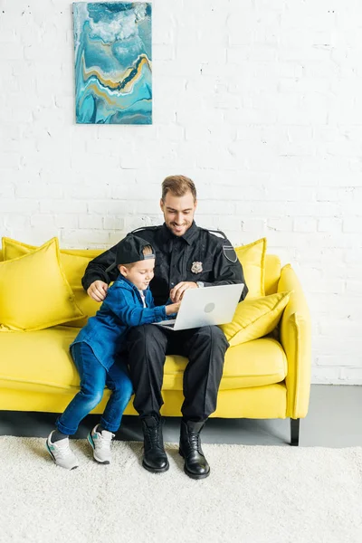 Guapo joven padre en uniforme de policía y el hijo usando el ordenador portátil juntos mientras están sentados en el sofá amarillo en casa - foto de stock