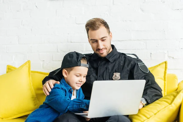 Beau jeune père en uniforme de police et fils utilisant ordinateur portable ensemble tout en étant assis sur le canapé jaune à la maison — Photo de stock