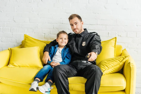 Padre joven en uniforme de policía viendo la televisión con su hijo mientras está sentado en el sofá amarillo en casa - foto de stock