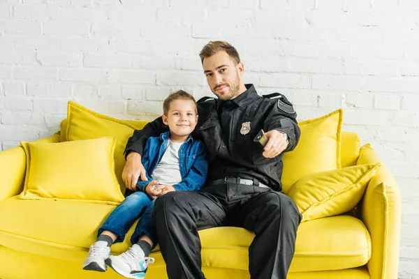 Schöner junger Vater in Polizeiuniform vor dem Fernseher mit Sohn, während er zu Hause auf der gelben Couch sitzt — Stockfoto