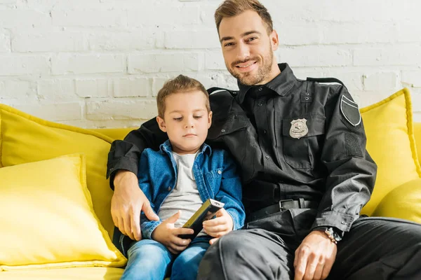 Heureux jeune père en uniforme de police regarder la télévision avec son fils assis sur le canapé jaune à la maison — Photo de stock