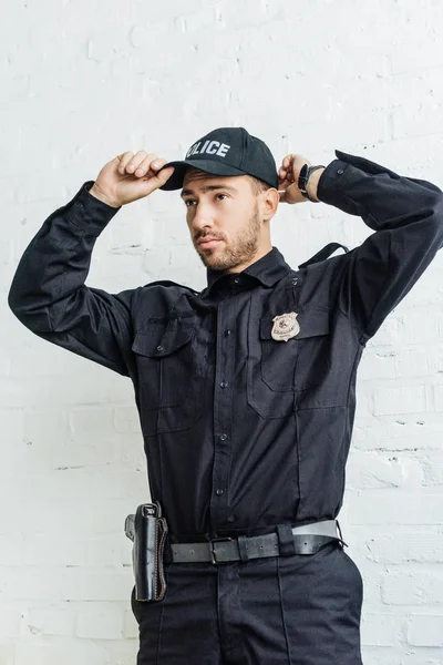 Handsome young policeman putting on cap in front of white brick wall — Stock Photo