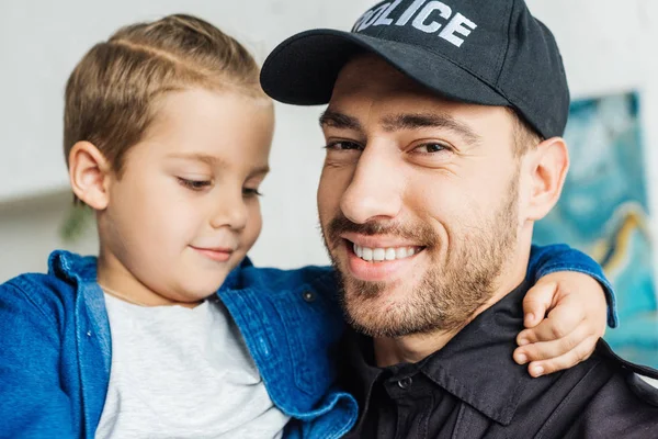 Close-up retrato de feliz jovem pai em uniforme policial carregando seu filhinho e olhando para a câmera — Fotografia de Stock
