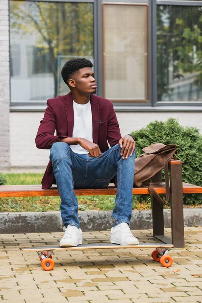 Schöner junger Mann mit Lederrucksack und Skateboard sitzt auf Bank auf der Straße — Stockfoto