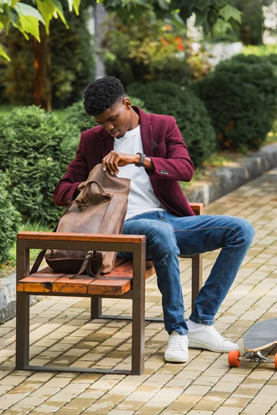 Jovem estudante elegante com mochila de abertura de skate enquanto sentado no banco na rua — Fotografia de Stock
