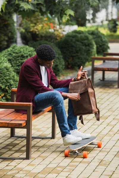 Hübsche junge Studentin mit Skateboard öffnet Rucksack, während sie auf Bank auf der Straße sitzt — Stockfoto