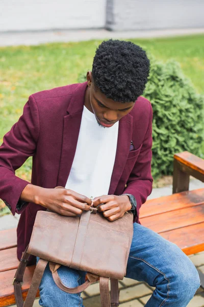 Handsome young student opening leather backpack while sitting on bench on street — Stock Photo