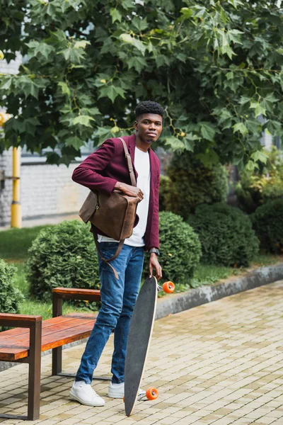 Красивый молодой студент с рюкзаком и скейтбордом, стоящий на улице — стоковое фото