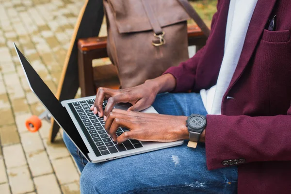 Plan recadré de pigiste afro-américain en utilisant un ordinateur portable sur banc avec sac à dos en cuir et planche à roulettes — Photo de stock